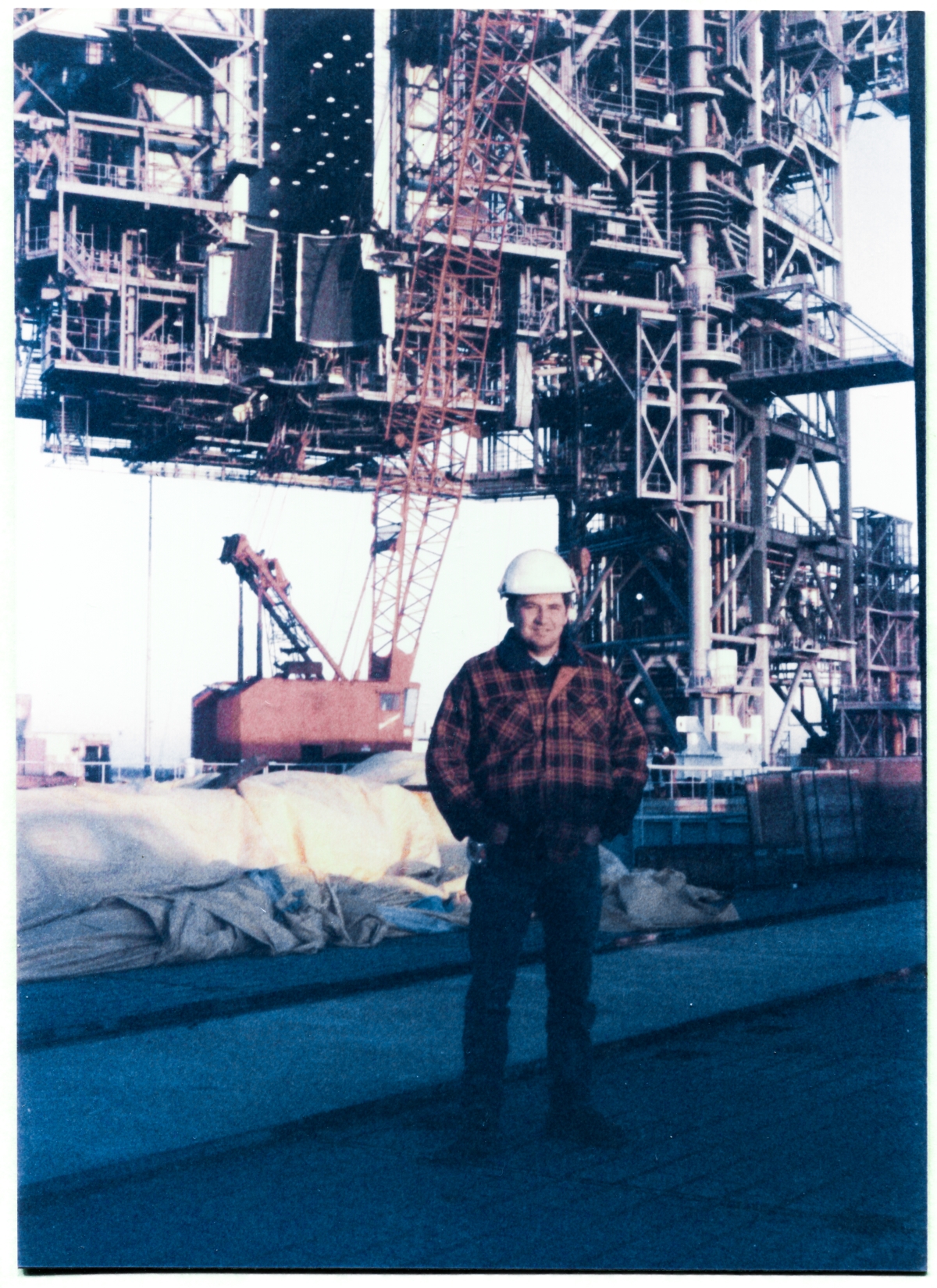 Image 139. Steve Skinner, Union Ironworker foreman working for Ivey Steel at the time, stands on the East side of the Flame Trench at Space Shuttle Launch Complex 39-B, Kennedy Space Center, Florida, with the lower portions of the Rotating Service Structure and the Fixed Service Structure in the distance behind him. Mostly blocked from view, down on the ground, other Union Ironworkers are preparing the Payload Ground Handling Mechanism (PGHM) Bridge Beam for today's Lift. They will be placing it inside the Payload Changeout Room up on a set of heavy Rails, which will allow it to roll forwards and backwards inside the PCR, just beneath its ceiling. The Bridge Beam supports the rest of the PGHM (which was installed later) on a set of quite-large hinge-pins, hanging down beneath the Bridge Beam all the way to where you can step right off of it, onto the PCR Main Floor, which it contacts via a set of small guide rails, but is in no way supported by them. The PGHM was an incredible thing, possessed of capabilities that defy belief and even imagination, and the Bridge Beam that rigidly held it all up was very large, very sturdy, and very heavy. How Wade Ivey and Rink Chiles ever managed to work out their beyond-cunning procedure for lifting it up onto those rails, up in the very topmost reaches of the PCR the way they did, still leaves me wondering in awe-struck amazement, and this Lift was probably the most radical thing of any sort that I ever witnessed being done during my entire ten years working structural steel at Cape Canaveral Air Force Station (now Cape Canaveral Space Force Station) and the Kennedy Space Center. To call this lift a ”tour de force” is to damn it with faint praise to an unforgivable degree. Photo by James MacLaren.